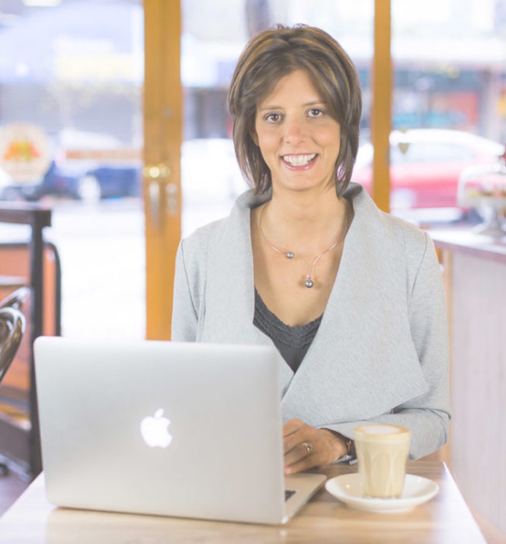 Dora Denk smiling at a café table with her laptop and a coffee